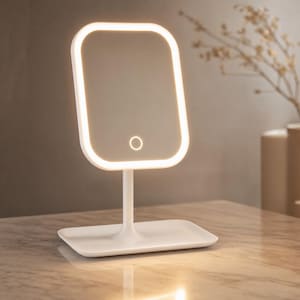 Puede incluir: Un espejo de tocador rectangular blanco con esquinas redondeadas y una tira de luz LED integrada alrededor del borde. El espejo está montado sobre un soporte vertical delgado unido a una bandeja base rectangular plana con un botón táctil.
