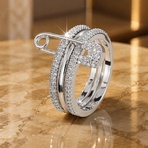 Puede incluir: Un anillo de tono plateado con forma de imperdible, que presenta una banda incrustada con numerosas piedras pequeñas, claras y brillantes. El diseño imita la forma de un imperdible clásico, con la cabeza y el cierre también cubiertos de piedras.