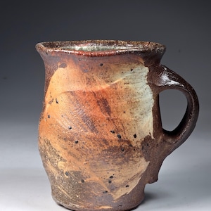 May include: A handcrafted ceramic mug with a textured, uneven surface featuring a blend of earthy brown, tan, and burnt orange tones. The mug has a wide, flared rim, a thick curved handle, and a mottled, speckled finish throughout the exterior.