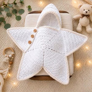 May include: A white star-shaped crochet swaddle blanket featuring a hood and three small brown wooden buttons along one side. The star design has five points and is edged with a light beige trim, providing a textured appearance for this baby item.