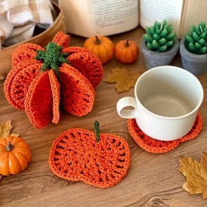 Puede incluir: Una calabaza naranja tejida a ganchillo con tallo verde, un posavasos plano con forma de calabaza y una taza de cerámica blanca sobre un segundo posavasos naranja. El conjunto presenta hilo naranja vibrante y detalles en verde tejido.