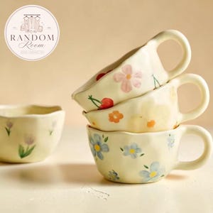 May include: A stack of three cream-coloured ceramic mugs with irregular, wavy rims and hand-painted floral and cherry motifs, accompanied by a single matching bowl featuring small purple tulip illustrations on the side.