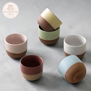 May include: A collection of six small ceramic cups with a two-tone design. Each cup features a dark brown base and a speckled pastel upper section in colors including pink, light green, yellow, white, and light blue. The cups are handleless.
