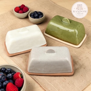 Può includere: Tre burriere rettangolari in ceramica con coperchio, che includono una versione bianca solida, una verde oliva e una grigio chiaro maculata, ciascuna poggiante su una base color terracotta con una piccola maniglia superiore.