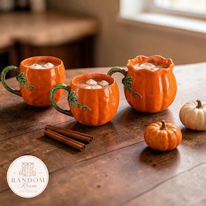 Peut inclure: Trois tasses en céramique orange en forme de citrouille, chacune dotée d'une anse en forme de tige verte et remplie d'une boisson garnie de guimauves. Deux petites citrouilles décoratives et trois bâtons de cannelle sont posés sur la table.