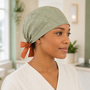 Puede incluir: Un gorro quirúrgico de color verde salvia con un delicado estampado floral blanco. El gorro presenta una corona ajustada e incluye una cinta de tela de color naranja quemado en la parte posterior, anudada en un lazo para asegurar el ajuste.