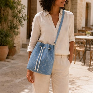 Peut inclure: Un sac seau en jean bleu clair orné d'un délicat motif floral blanc, doté d'une longue bandoulière réglable et d'une fermeture à cordon blanc avec des extrémités nouées. Le sac est porté en bandoulière, révélant une forme cylindrique structurée.