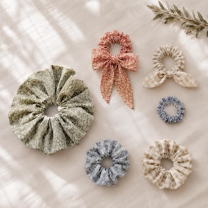 May include: A collection of six fabric hair scrunchies in various sizes and patterns. Included are a large sage green floral scrunchie, a terracotta floral scrunchie with long bow tails, a beige gingham scrunchie with bunny ears, and three smaller floral or gingham scrunchies in blue and beige tones.