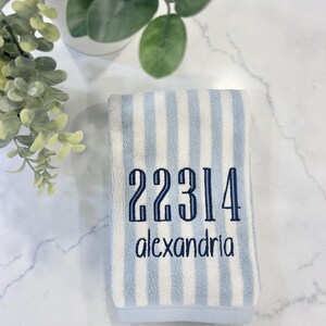 May include: A rectangular hand towel featuring vertical light blue and white stripes. The towel is embroidered with the dark blue numbers 22314 and the word alexandria in a lowercase sans-serif font centered on the striped fabric surface.