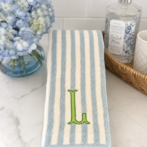 May include: A hand towel featuring vertical light blue and cream stripes with a lime green embroidered letter L centered on the front. The towel has a finished hem at the bottom and a soft, textured fabric surface suitable for bathroom use.