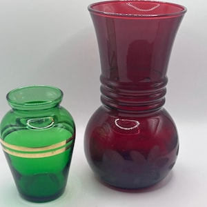 May include: A small green glass vase with two horizontal gold bands around the centre and a larger, taller ruby red glass vase with a bulbous base, a ribbed neck, and a flared rim, both featuring smooth, translucent surfaces and reflective highlights.