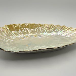 May include: An oval-shaped ceramic dish featuring a scalloped, shell-like edge and a lustrous, iridescent finish. The surface displays a radial ribbed texture with a pearlescent sheen that shifts between soft gold, cream, and pale blue tones.