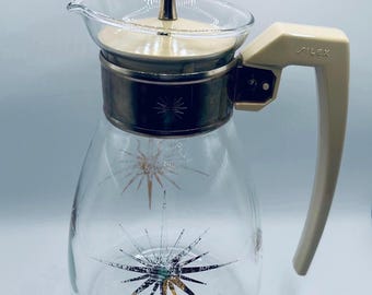 May include: A clear glass carafe featuring a starburst pattern in gold and black. The carafe has a cream-colored plastic handle with the word Silex embossed on it, a matching cream lid with a gold-toned finial, and a metallic band around the neck.