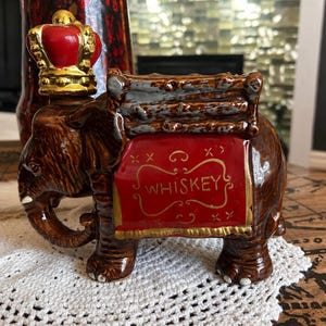 May include: A brown ceramic elephant decanter featuring a red and gold blanket with the word WHISKEY printed in gold. The elephant wears a gold crown with a red top, and a brown textured saddle sits on its back with grey highlights.