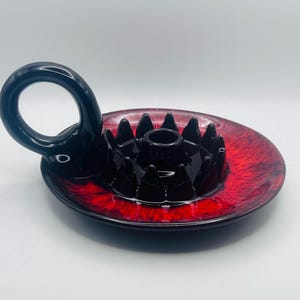 Peut inclure: Un bougeoir en céramique noir et rouge avec une base circulaire au centre rouge vif et aux bords noirs. Le centre présente une coupelle à bougie surélevée entourée d'une couronne de pointes, avec une grande anse circulaire sur le côté.