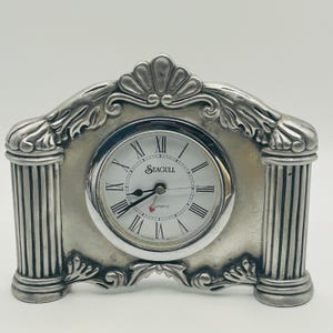 May include: A silver-toned metal mantel clock featuring a white circular face with black Roman numerals and the text Seagull Quartz. The clock is framed by two fluted columns and a decorative arched top with a shell-like motif at the center.