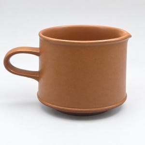 Peut inclure: Une tasse en céramique de couleur terre cuite unie avec une finition mate lisse. La tasse présente un corps cylindrique avec un bord légèrement évasé, un petit bec verseur intégré sur le côté droit et une anse incurvée sur le côté gauche.