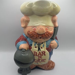 May include: A ceramic pig figurine dressed as a chef, wearing a white toque and apron with the text BAR B QUE PIG-OUT. The pig has a brown mustache, a red shirt, and blue overalls, holding a dark gray round grill and a black spatula in hand.
