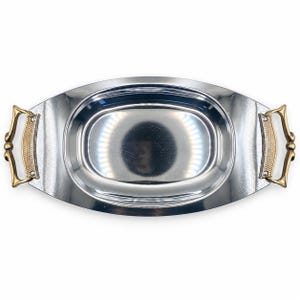 May include: An oval-shaped serving tray with a polished silver-tone finish and a smooth, reflective surface. The tray features two decorative gold-tone metal handles on each side, each with a textured central panel and a curved, ornate frame.