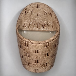 May include: A wall-mounted ceramic pocket with a light brown, textured finish that mimics the appearance of woven wicker. The piece features a semi-circular opening at the top and a rounded base, with a smooth, off-white interior surface.