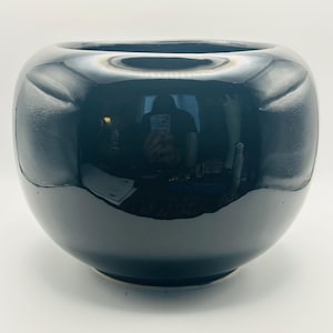 May include: A glossy black ceramic bowl with a rounded, bulbous shape and a wide, open top. The surface features a smooth, reflective finish with subtle, textured indentations on the sides, creating a distinct contrast against the dark glaze.