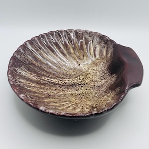 May include: A ceramic bowl shaped like a seashell with a glossy finish. The interior features a mottled pattern of deep burgundy, cream, and golden brown tones, while the exterior rim is a solid, dark, polished burgundy with a small handle.