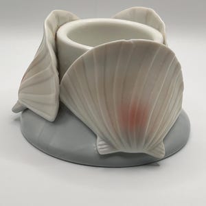 May include: A white ceramic egg cup featuring a central cylindrical holder surrounded by three sculpted scallop shells. The shells are off-white with subtle pinkish-orange accents at the base, all mounted on a smooth, circular light grey base.