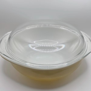 May include: A round, light yellow glass casserole dish with integrated side handles and a clear, domed glass lid. The base of the dish features a smooth, opaque finish, while the lid is transparent, allowing for a clear view of the interior.