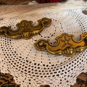 May include: Two matching brass drawer pulls featuring a curved, ornate shape with a central ball detail and an engraved linear knot pattern. Each handle has two mounting points on the ends, showcasing a polished metallic finish with dark accents.