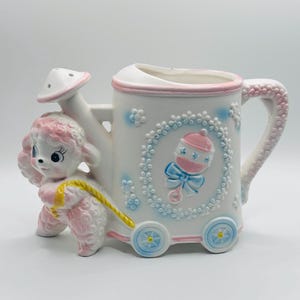 May include: A white ceramic watering can planter featuring a pink poodle figure on the front. The body is decorated with a raised blue floral wreath surrounding a pink and blue baby rattle. The handle and base are accented with pink and blue.