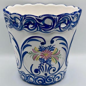 May include: A white ceramic planter featuring a scalloped rim and a hand-painted blue scrollwork pattern. The centre of the vessel displays a floral motif in shades of pink, yellow, and green, framed by intricate blue swirling lines and borders.