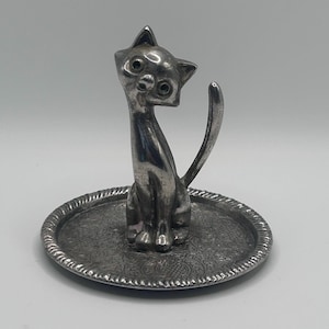 May include: A silver-toned metal ring holder featuring a stylised cat figure sitting upright in the centre of a circular tray. The tray has a textured, ornate pattern on its surface and a rope-like border detail around the outer edge.