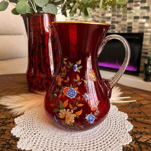 May include: A deep red glass jug featuring a hand-painted floral design with blue, orange, and yellow blossoms, green leaves, and a butterfly. The jug has a clear glass handle and a gold-toned rim, resting on a white crocheted doily.