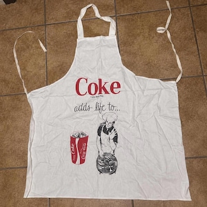May include: A white apron featuring the red text Coke and the phrase adds life to... in black. The graphic includes a red cup with ice and a black and white illustration of a chef holding a platter of food. It has white fabric neck and waist ties.