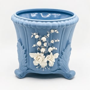 May include: A light blue planter featuring white relief floral decorations including roses and lily of the valley. The vessel has a flared rim with a beaded edge, vertical fluted panels, and stands on three ornate feet with leaf-shaped accents.
