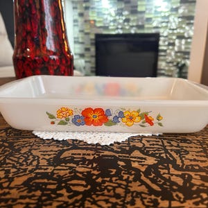 May include: A rectangular white glass baking dish featuring a colourful floral pattern on the side. The design includes orange, yellow, and blue flowers with green leaves. The dish has smooth, rounded edges and a glossy finish, presented as a shallow container.