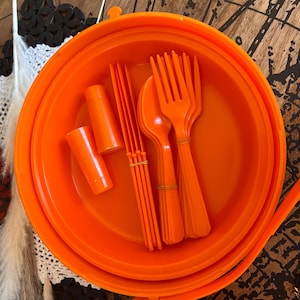 May include: A bright orange plastic bowl containing a set of matching orange plastic utensils, including several forks, spoons, and small cylindrical components, all neatly arranged and bundled together within the circular container.