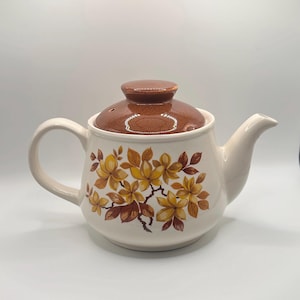May include: A cream-coloured ceramic teapot featuring a brown speckled lid and a floral pattern in shades of yellow, orange, and brown. The teapot has a curved handle, a short spout, and a rounded body, showcasing a botanical design on the front.