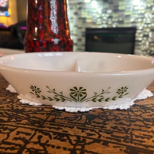 May include: A white oval divided serving dish made of milk glass featuring a green floral pattern of daisies and leafy vines along the exterior side. The dish has integrated handles on both ends and a central divider separating the interior space.