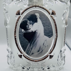 May include: A clear glass picture frame with an ornate, molded border and a central oval opening. The frame features a metallic, rope-textured inner trim with decorative accents at the top, bottom, and sides, holding a black and white wedding photo.