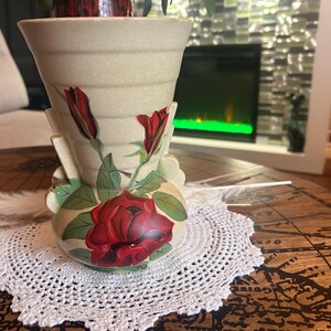 May include: A beige ceramic vase with a flared, ribbed rim and a rounded base. The vase features a hand-painted design of a large, blooming red rose at the bottom, accompanied by two smaller red rosebuds on green stems with leaves.
