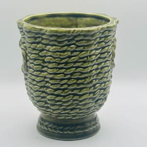 May include: A ceramic planter with a glossy olive green finish and a textured exterior that mimics the appearance of woven rope. The vessel features a scalloped rim and a circular pedestal base, with a consistent rope-like pattern throughout.
