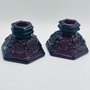 May include: A pair of deep purple glass candle holders featuring a hexagonal base and a raised diamond pattern. Each piece has a wide, flat bottom and a central circular opening at the top designed to securely hold a standard taper candle.