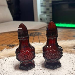 May include: A pair of deep red glass salt and pepper shakers featuring a pointed cap design and a textured, diamond-patterned body. The shakers have a distinct, moulded glass construction with a wide base and a tapered neck, showcasing a rich, translucent crimson hue throughout the glass material.