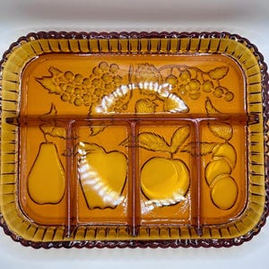 May include: A rectangular amber glass serving tray with three internal dividers. The tray features a moulded fruit motif including grapes, a pear, an apple, a peach, and berries. It has a scalloped edge and two side handles with three small circular accents.