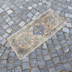 May include: A rectangular woven rug featuring a faded, muted color palette of dusty pink, sage green, and slate blue. The design consists of repeating geometric medallions and intricate floral motifs arranged in a symmetrical, traditional pattern.