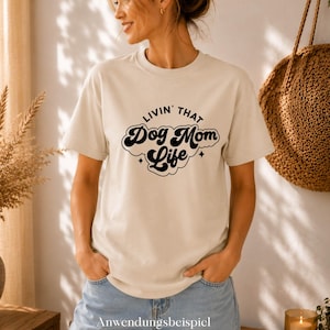 Può includere: Una t-shirt beige a maniche corte con la scritta nera "LIVIN' THAT Dog Mom Life" in un carattere stilizzato e ondulato. Il design include piccoli accenti a forma di stella a quattro punte sopra e sotto il testo, centrato sul petto.