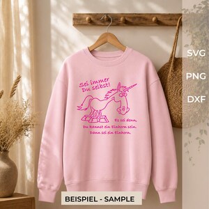 Puede incluir: Una sudadera de manga larga de color rosa claro con un gr&aacute;fico de color rosa oscuro de un unicornio de dibujos animados. El texto dice: Sei immer Du selbst! Es sei denn, Du kannst ein Einhorn sein. Dann sei ein Einhorn. La prenda est&aacute; colgada.
