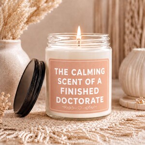 May include: A clear glass candle jar with a lit wick and a black metal lid leaning against the side. The label is a muted peach color with white text that reads THE CALMING SCENT OF A FINISHED DOCTORATE, accented by small leaf and heart graphics.