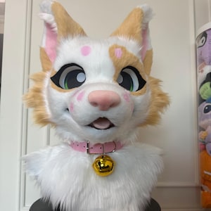 Op de afbeelding: Een dierenkopmasker van wit en beige imitatiebont met grote expressieve ogen met zwarte omlijning, een roze neus en roze vlekken op het voorhoofd en de wangen. Het masker heeft een lichtroze halsband met een grote, glanzende goudkleurige metalen bel.